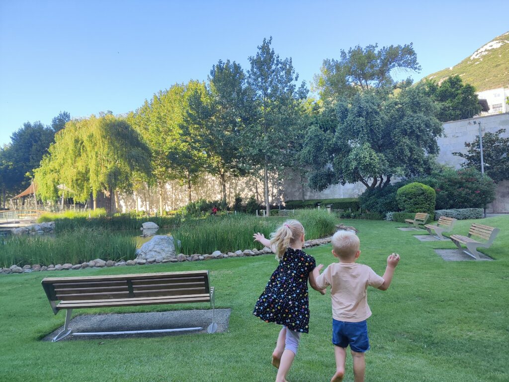 Dwoje dzieci bawi się na trawie w Commonwealth Park w Gibraltarze, w otoczeniu zieleni i stawu
